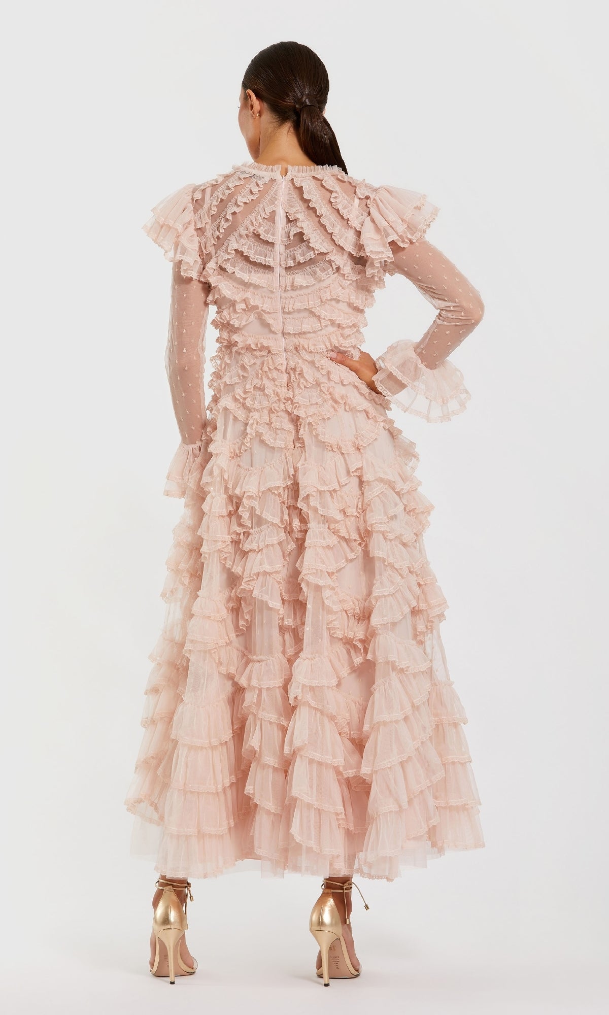 A woman faces away, wearing the Mac Duggal 8274 Long Sleeve Ruffle Formal Dress in pink with sheer sleeves and gold high heels, set against a plain white background.